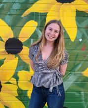 Emilia O'Brien, smiling, long dark blonde hair, jeans and a checked wrap top, standing against a wall painted with giant sunflowers